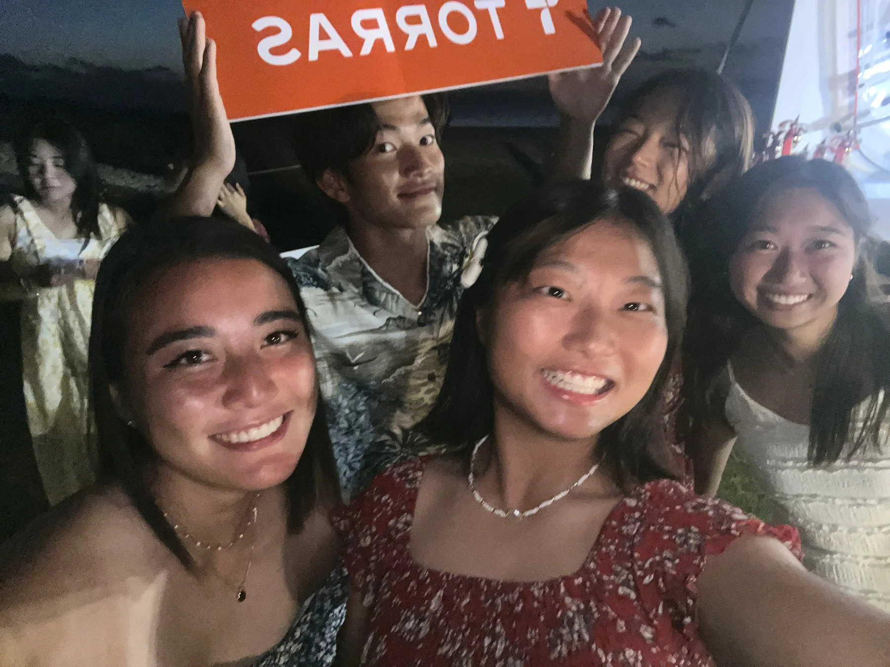 夜の屋外で、数人の若者が楽しそうに笑顔で写っているグループ写真です。背景には「TORRAS」と書かれた看板が見えます。