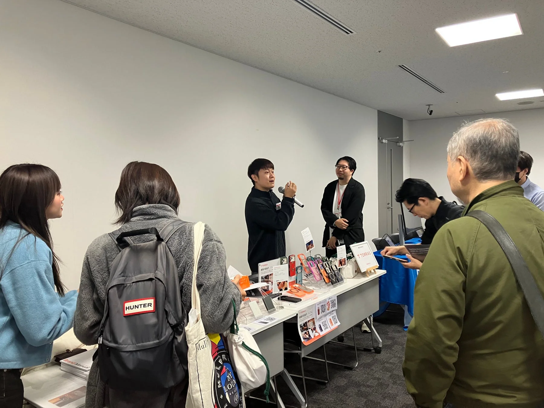 屋内のイベントで、男性がマイクを手に製品を説明しています。テーブルにはカラフルなスマートフォンアクセサリーが並び、来場者が熱心に見ています。