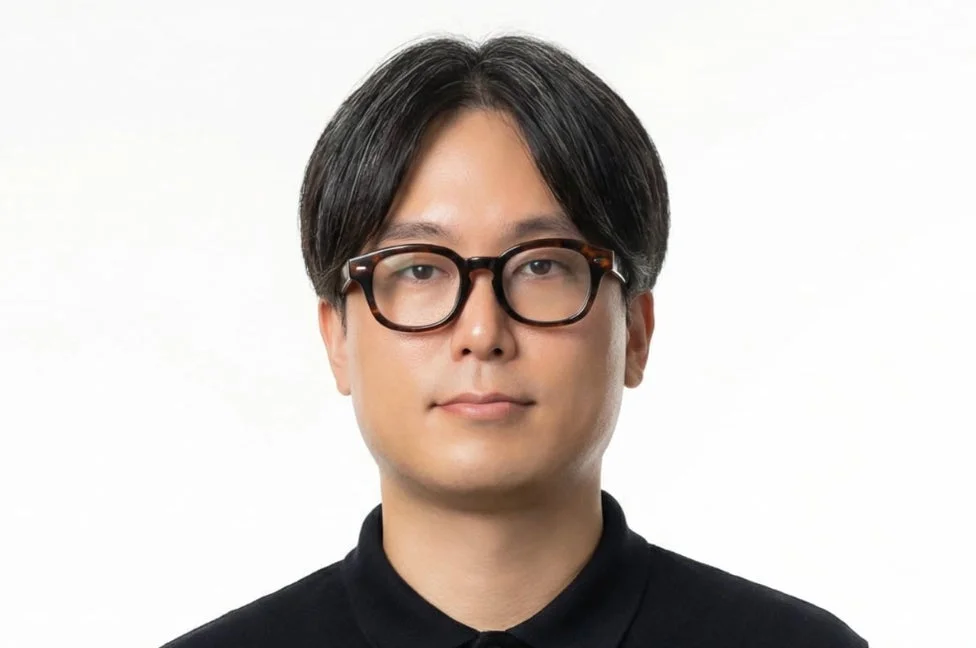 黒いポロシャツとべっ甲柄の眼鏡をかけたアジア系の男性が正面を向いて立っているポートレート写真