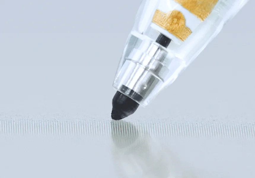 黒いマーカーペンのペン先が、わずかに凹凸のある白い紙のような表面に触れている様子をクローズアップで捉えた画像です。