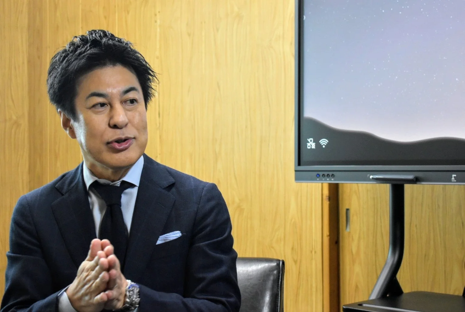 ダークスーツを着たアジア人男性が、木目調の壁と大型モニターの前で手を組みながら話しています