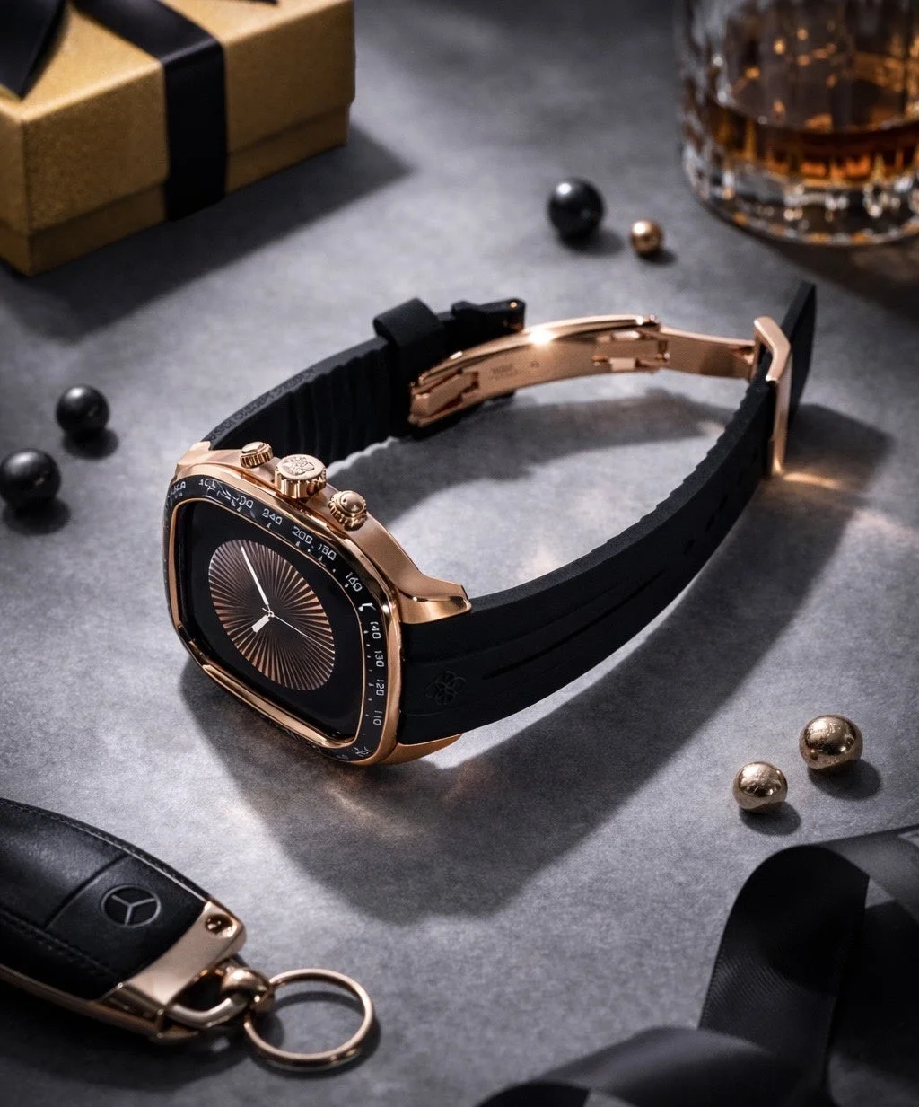 ローズゴールドの高級スマートウォッチが中心に置かれ、メルセデスベンツのキー、ウイスキーグラス、ギフトボックスが並ぶ洗練された構図。
