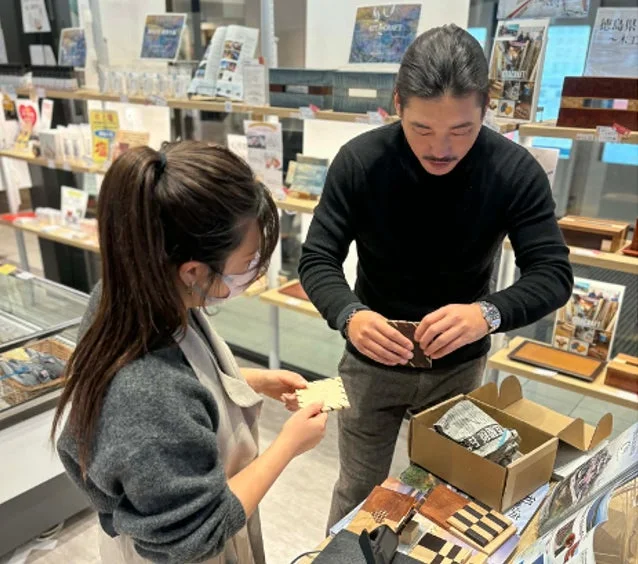 店舗で、男性客が女性店員から木工品の説明を受けている様子