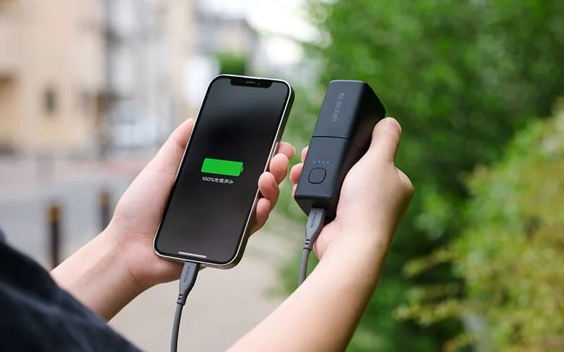 屋外でスマートフォンとモバイルバッテリーを手に持ち、充電している様子です。スマートフォンの画面には「100%充電済み」と表示されており、充電が完了していることが示されています。