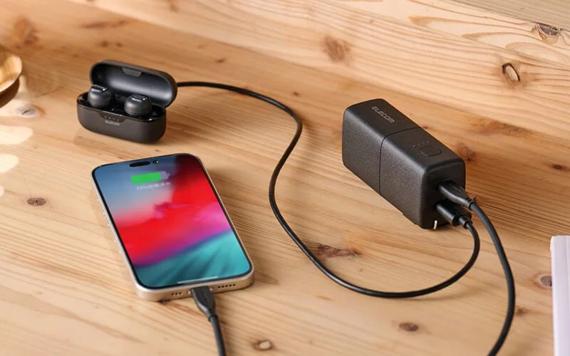 木目のデスク上で、スマートフォンとワイヤレスイヤホンが黒い充電器に接続され、同時に充電されている様子を捉えた画像です。現代の電子機器の充電風景を示しています。