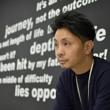 黒い背景に白い英文が書かれた壁の前で真剣な表情を浮かべる鈴木 教郎