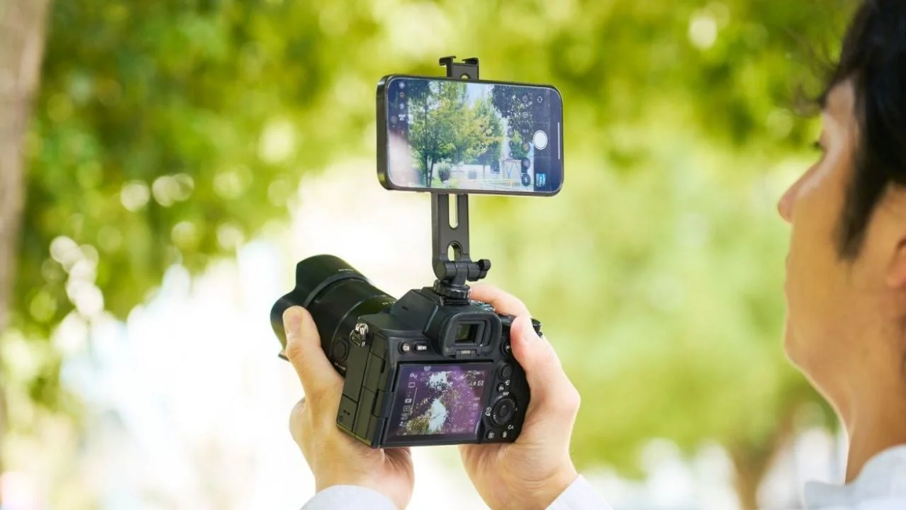 男性が一眼レフカメラにスマートフォンを装着して撮影しているシーン。緑豊かな屋外で、最新の撮影機材を組み合わせて使っている様子が伺えます。