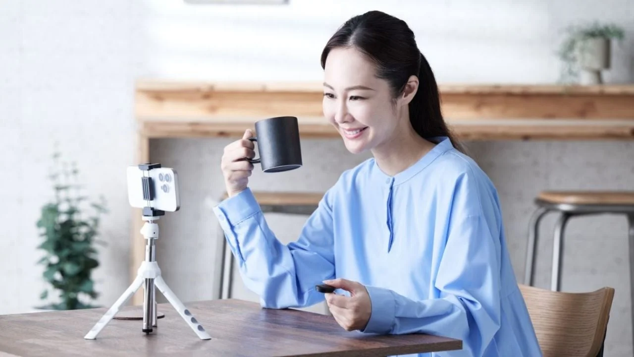 笑顔の女性が自宅のような場所で、スマートフォンを三脚にセットしてライブ配信やビデオ通話をしている様子です。コーヒーカップを片手にリラックスした雰囲気で、カジュアルな水色のシャツを着ています。