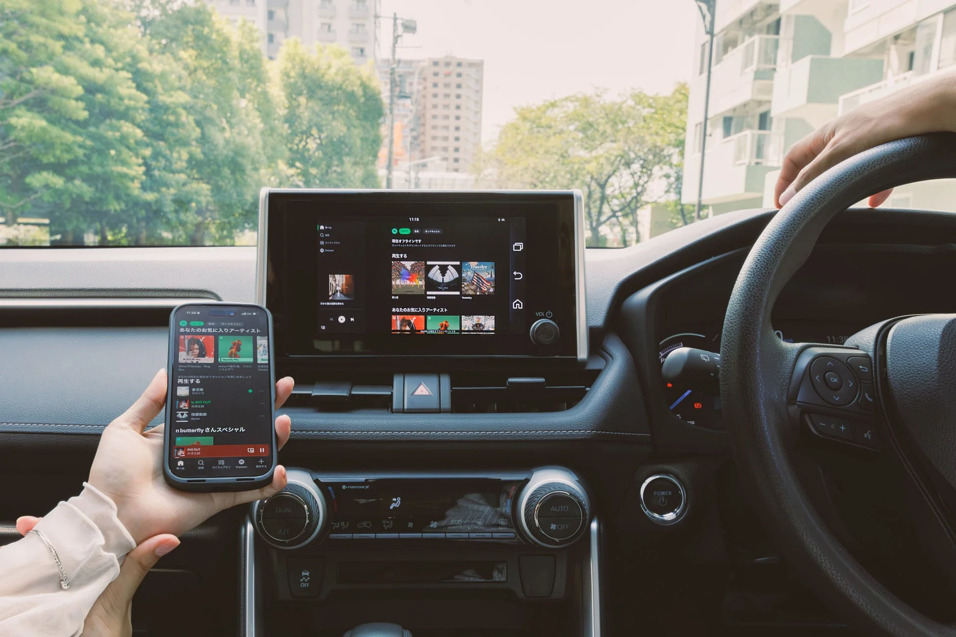 車内で撮影された写真で、ダッシュボード、カーナビ画面、そしてスマートフォンを持つ手が写っています。カーナビとスマートフォンには音楽アプリの画面が表示されており、運転中または停車中に音楽やポッドキャストを楽しんでいる様子が伺えます。背景には木々や建物が見えます。