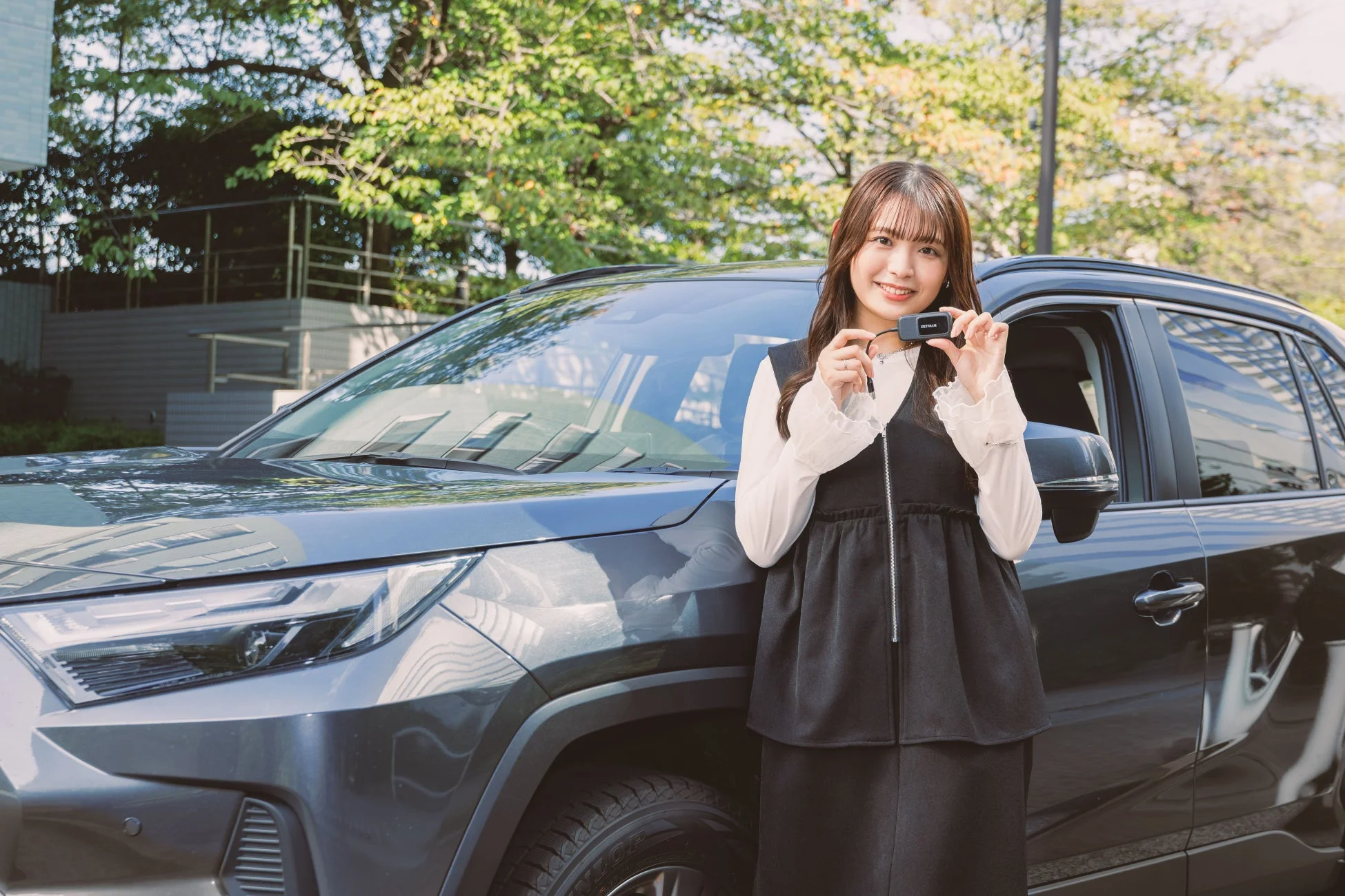 笑顔の若い女性がダークグレーのSUVの横に立ち、DETERIAと書かれた小型デバイスを両手で持って見せています。車と製品をアピールする様子です。
