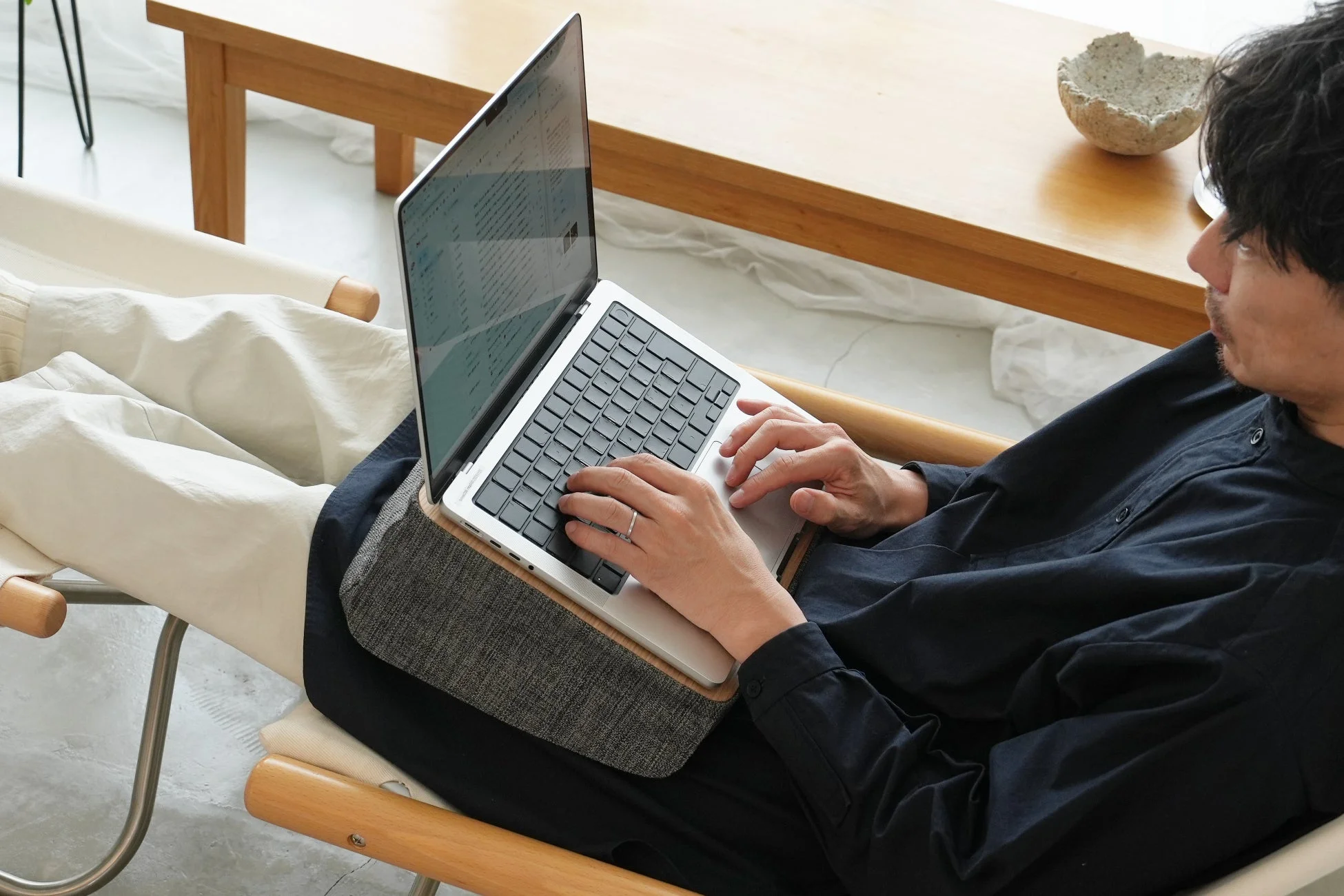 男性がリラックスした姿勢で椅子に座り、ラップデスクに置いたノートパソコンで作業している様子