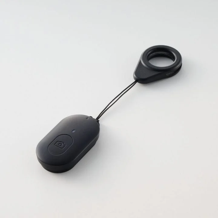 白い背景に置かれた黒いBluetoothシャッターリモコンです。カメラアイコンのボタンと指を通せるリング付きストラップが特徴で、スマートフォンでの写真撮影を遠隔で操作できます。