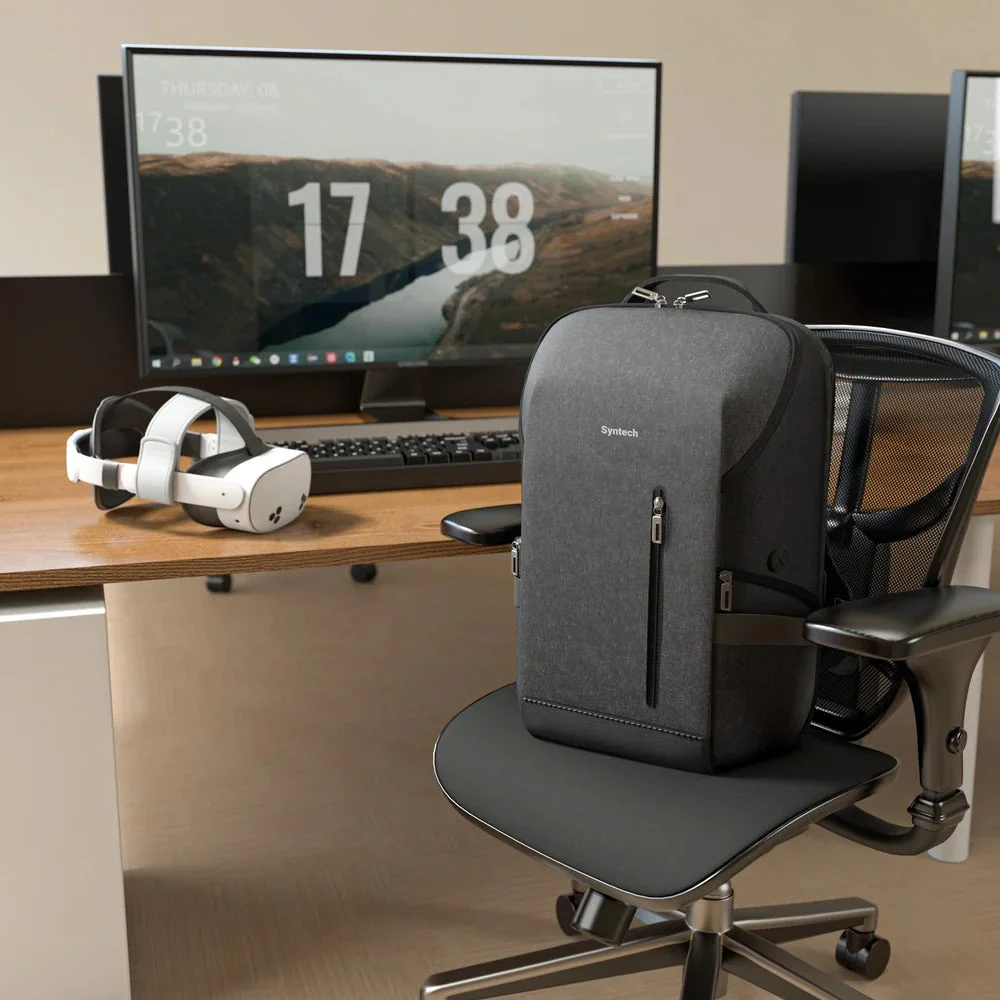 現代的なオフィス環境の一角を捉えた画像です。木製のデスクには白いVRヘッドセットが置かれ、その奥には時刻「17 38」と日付が表示された大型モニターがあります。手前のオフィスチェアには、Syntechのロゴが入ったグレーのバックパックが置かれています。