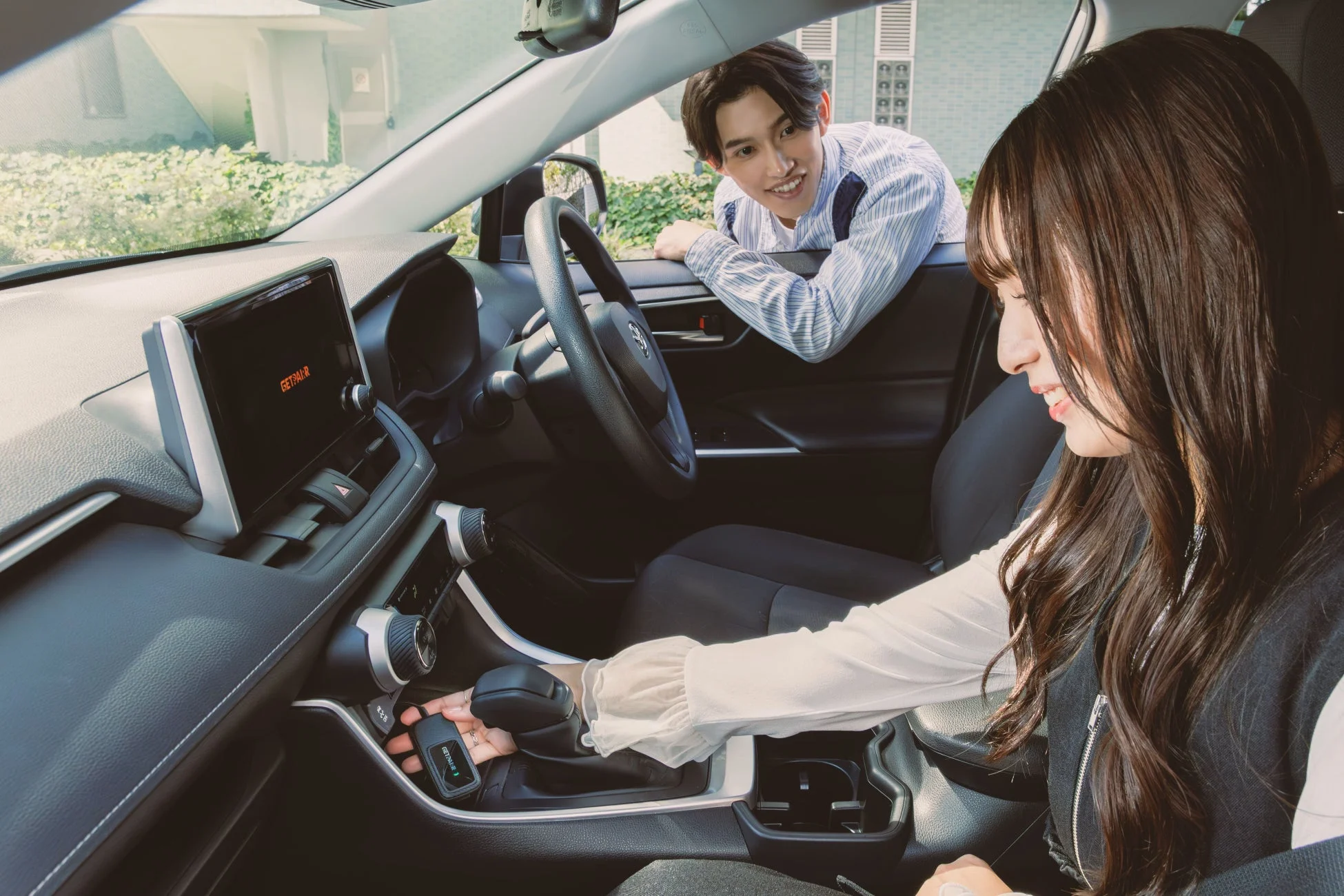 現代的な車内で、男性が運転席の窓から身を乗り出し、助手席の女性に笑顔で話しかけている。女性は手に「GETAIR」と書かれたデバイスを持ち、楽しげに微笑んでいる。和やかな交流が感じられる一枚。
