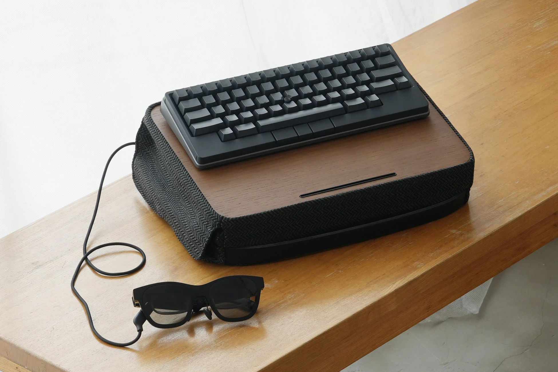 木製の台の上に置かれたミニマルな黒いキーボードと、その横にケーブルで繋がれたスマートグラスのセットアップ