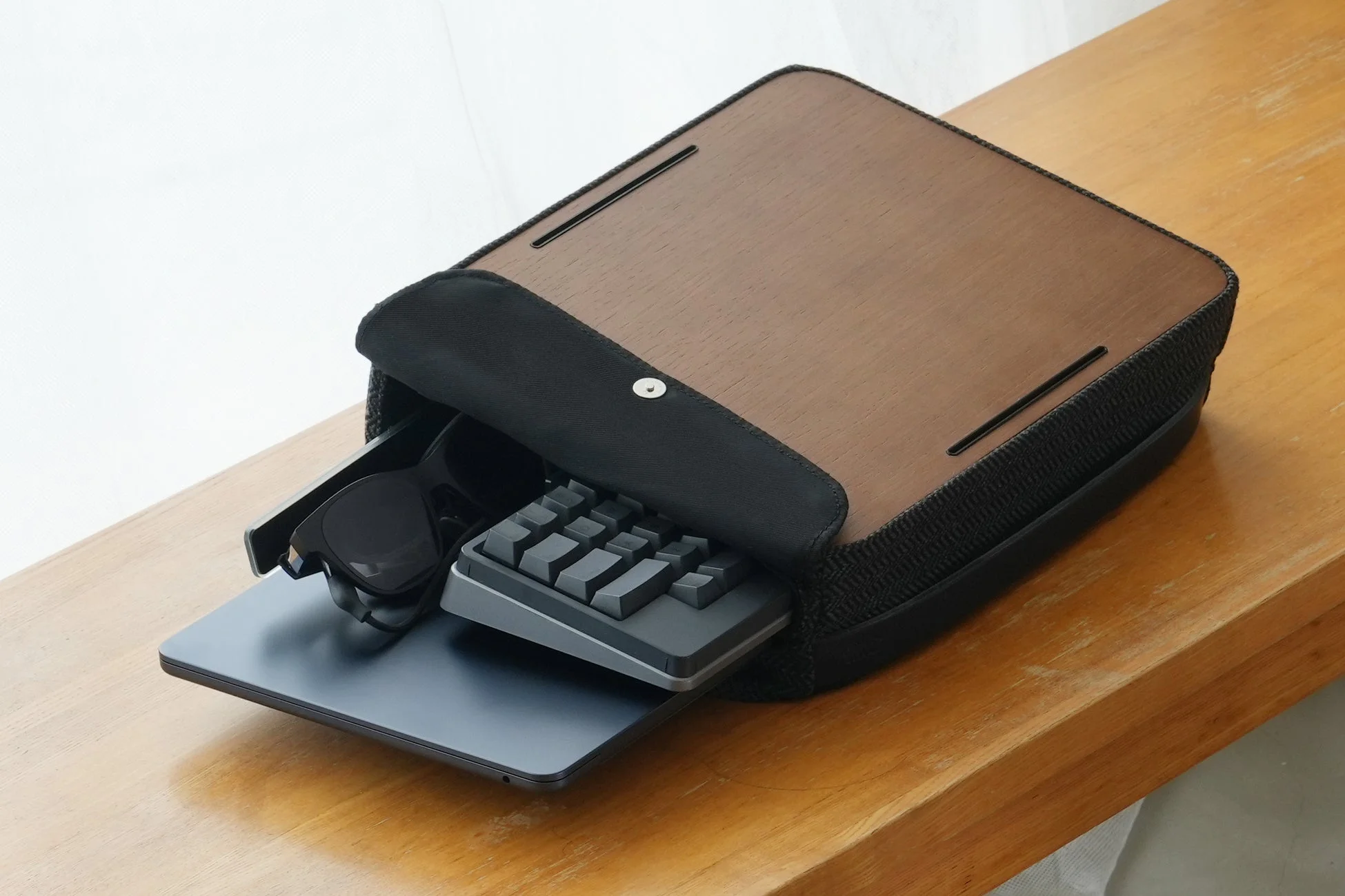 木製の天板を持つスタイリッシュなキャリングケースが木製のテーブルに置かれ、中からノートパソコン、コンパクトなキーボード、サングラスが見えている