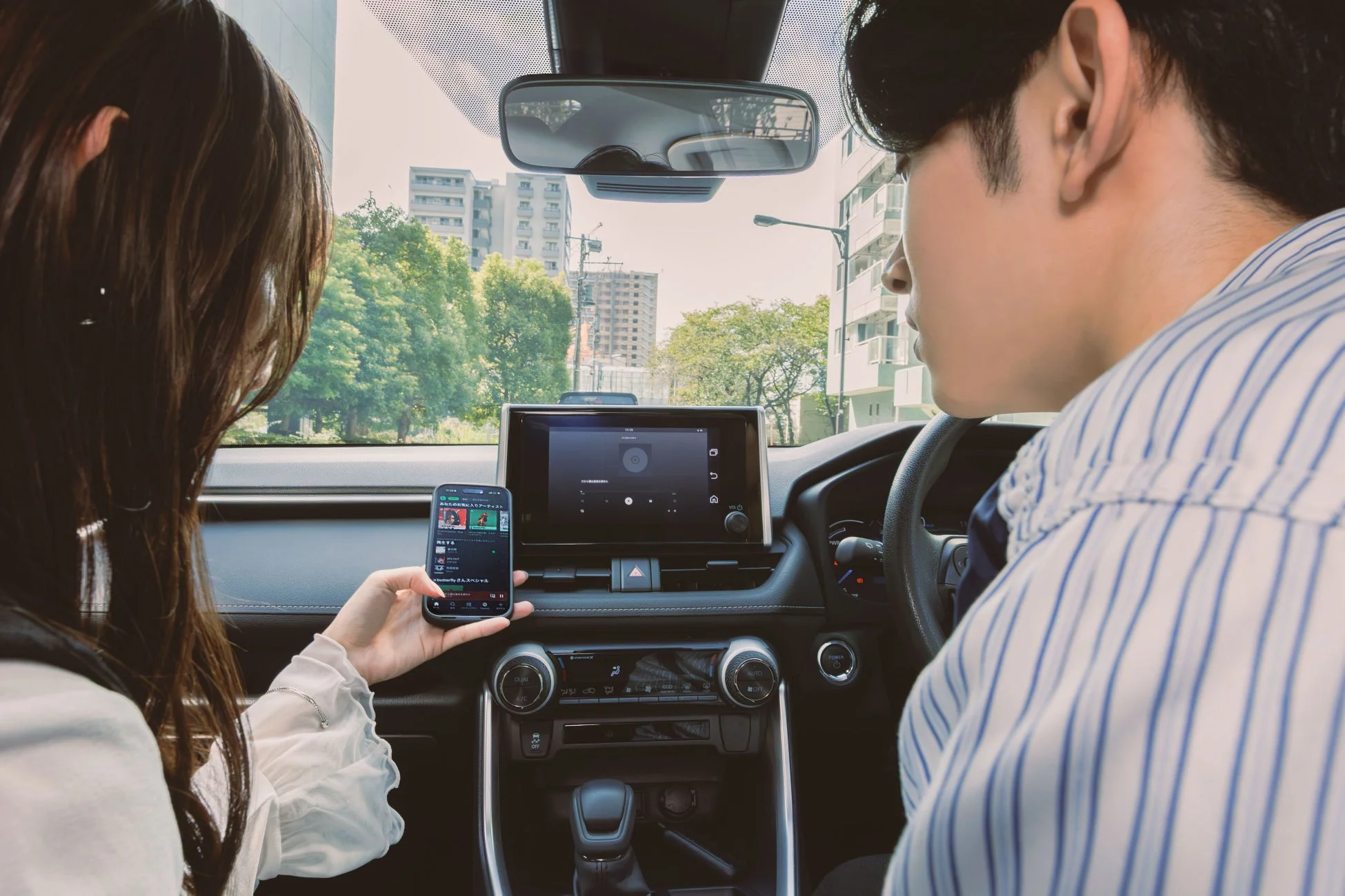 車内でくつろぐ男女のカップルが映っています。助手席の女性がスマートフォンを操作し、運転席の男性が前を向いています。カーナビ画面も確認でき、ドライブ中に音楽を選んでいる様子です。