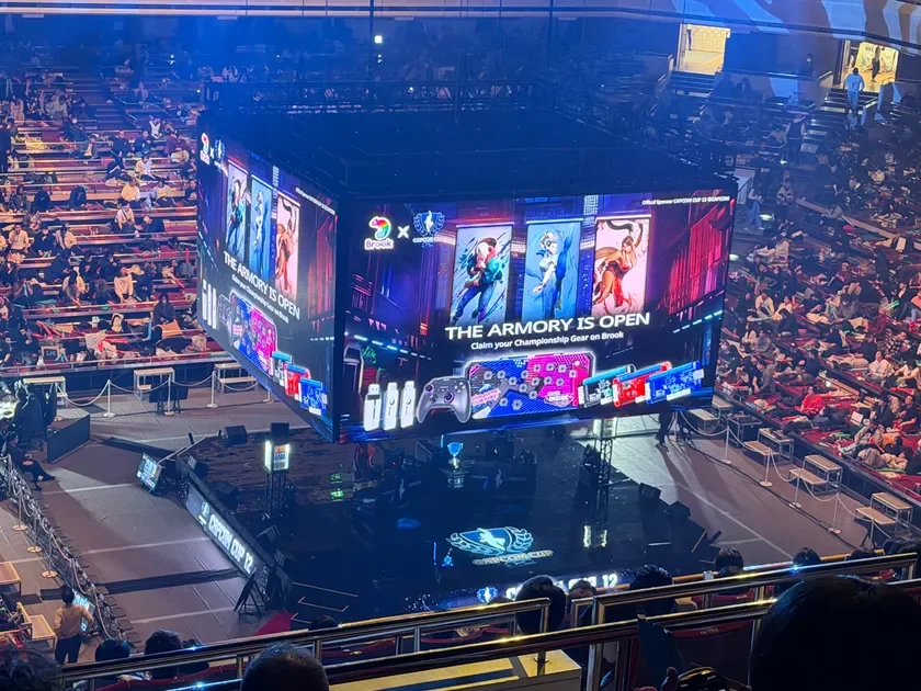 多数の観客で賑わう会場で、大型の四面スクリーンが中央に吊り下げられています。スクリーンには「CAPCOM CUP 12」のロゴや、ゲームキャラクター、ゲーミングデバイスの広告が表示されており、eスポーツイベントが開催されている様子です。