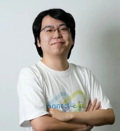 メガネをかけたアジア人男性が白いTシャツを着て腕を組み、カメラに向かって微笑んでいます。
