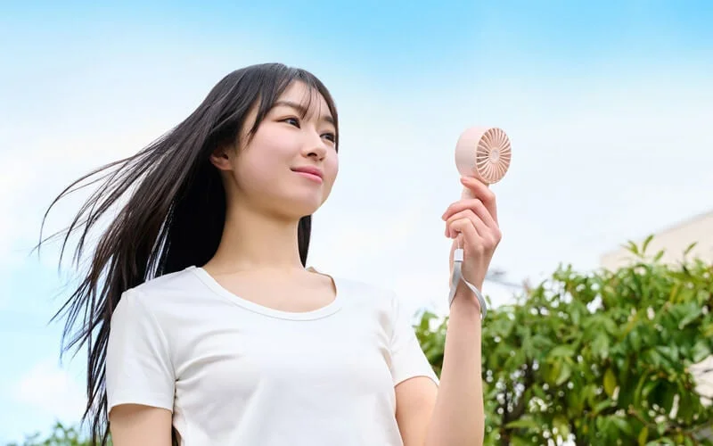 青空の下、屋外でハンディファンを使い涼んでいる女性