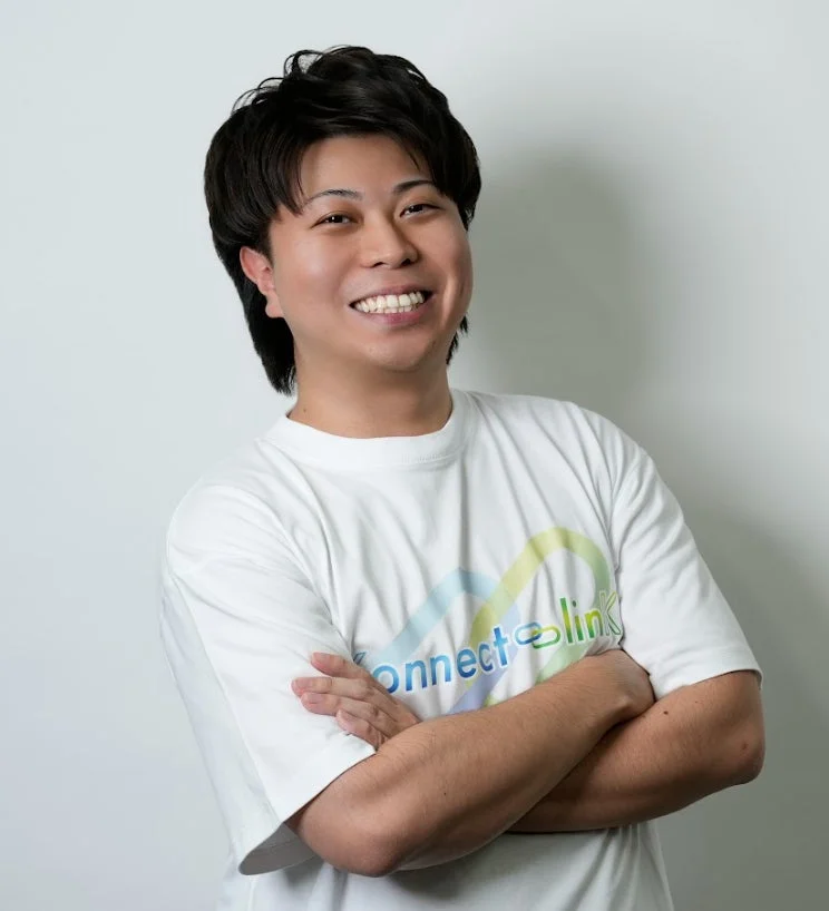 白いTシャツを着た若い男性がカメラに向かって満面の笑みを浮かべ、腕を組んで立っています。