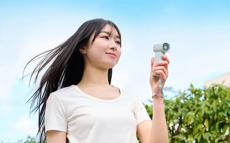 青空の下、白いTシャツを着た女性がポータブル扇風機を手に持ち、涼しげな表情