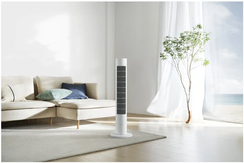 明るい日差しが差し込むリビングルームで、白いタワーファンが置かれています。ベージュのソファと観葉植物があり、窓の外には広大な海と空が広がる開放的な空間です。