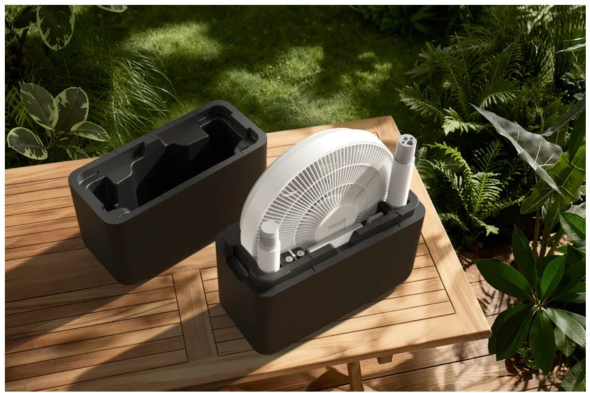 木製のテーブルの上に、折りたたみ式の白い扇風機が黒い収納ケースに収まっています。隣にはもう一つの空の収納ケースがあり、背景には緑豊かな植物が広がっています。