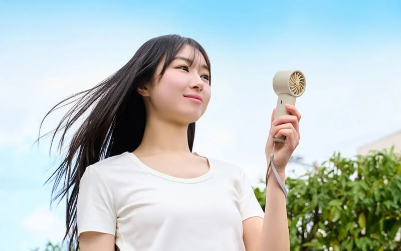 青空の下、白いTシャツを着た若い女性がハンディファンを持ち、涼んでいる様子