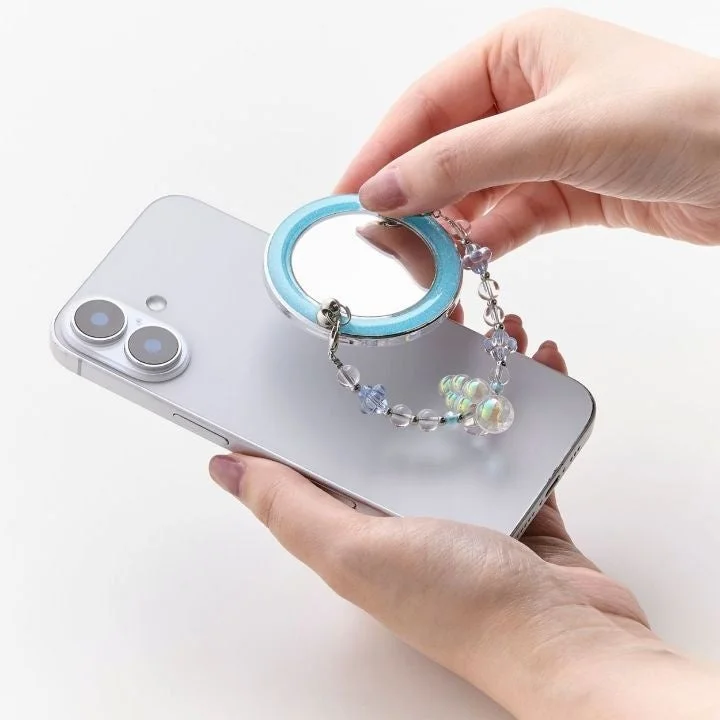 人物がシルバーのスマートフォンを手に持っており、背面には水色の縁取りとビーズのストラップが付いた丸いミラー型アクセサリーが装着されています