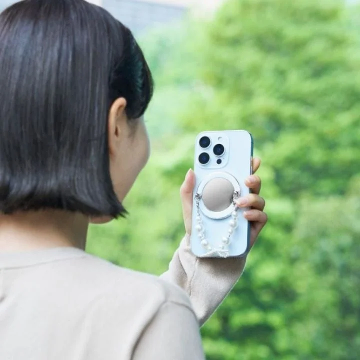 女性がパールストラップとミラー付きのライトブルーのスマートフォンを持っている後ろ姿です。背景には緑豊かな木々が見えます。