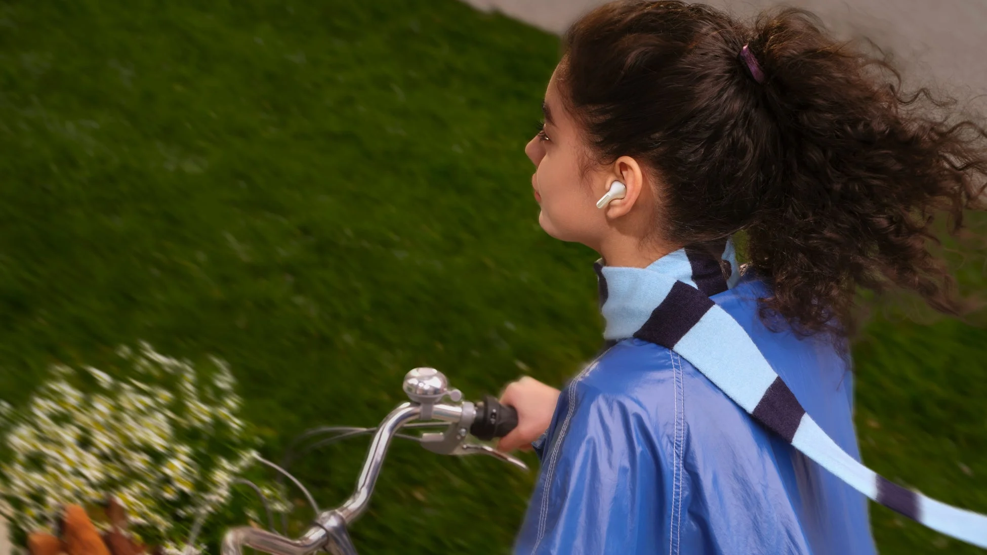 自転車に乗る若い女性が、風になびく髪とマフラーを身につけ、イヤホンで音楽を聴きながらサイクリングを楽しんでいる様子。背景には緑の草地が広がっています。
