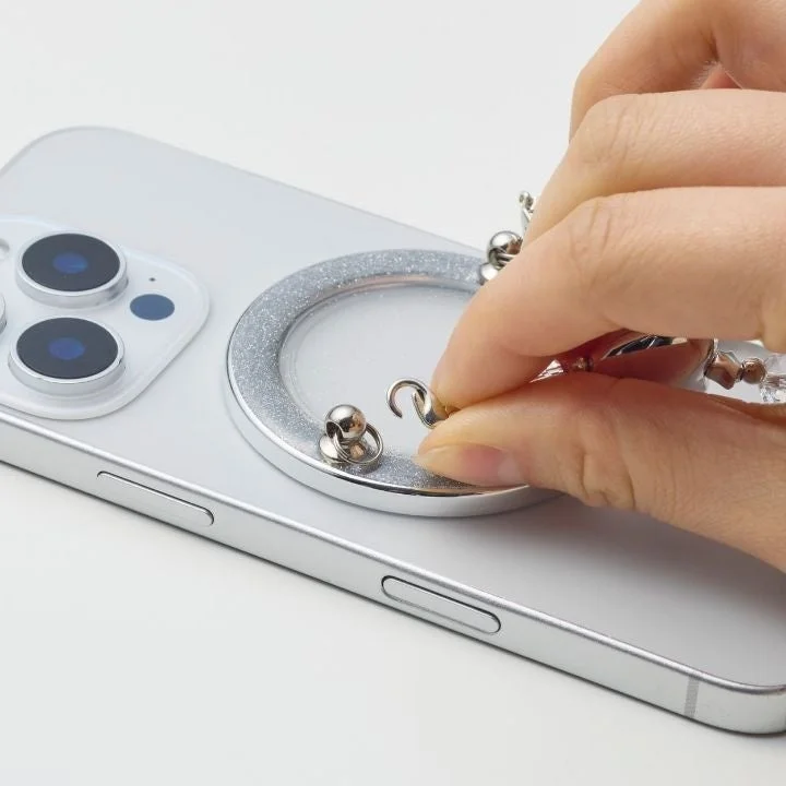 グリッター加工されたシルバーのリング型アタッチメントが付いたスマートフォンの背面に、手が小さなシルバーのチャームを取り付けている様子