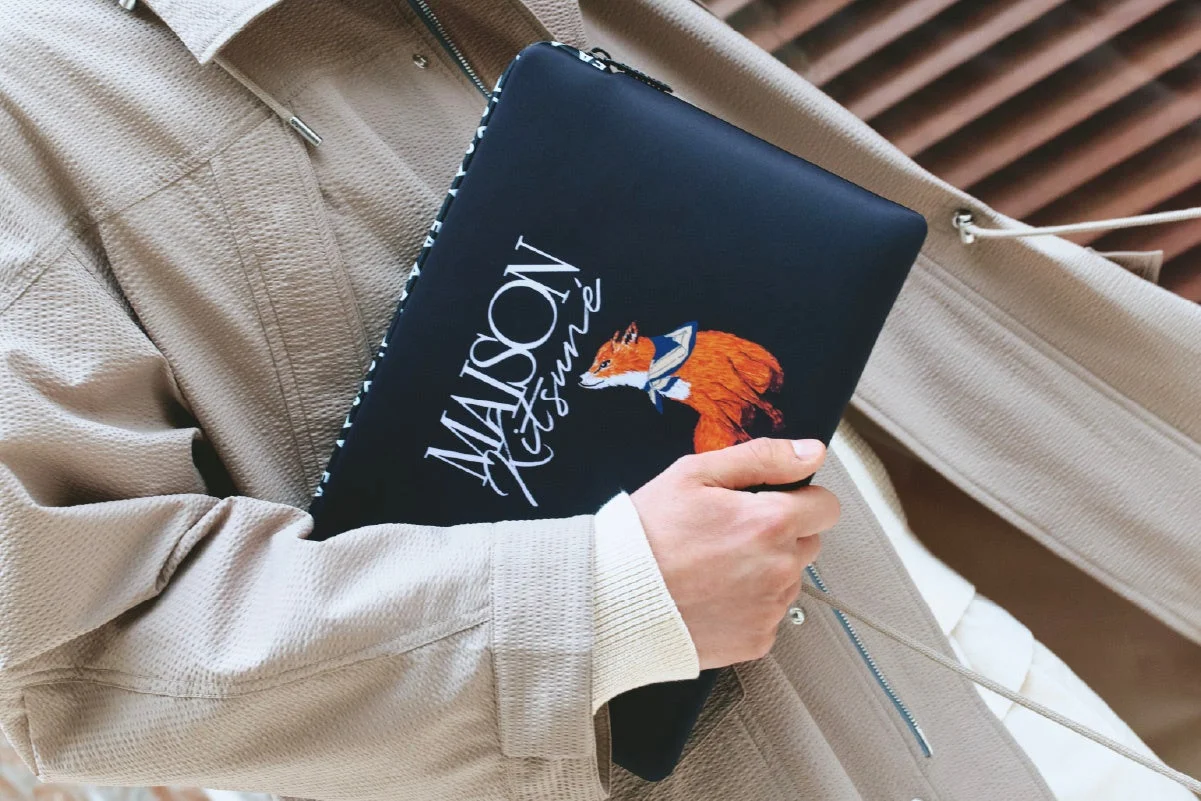 ベージュのジャケットを着た人物が、黒いメゾンキツネのクラッチバッグまたはPCケースを持っている様子。バッグにはブランドロゴとセーラー服を着たキツネのイラストが描かれています。