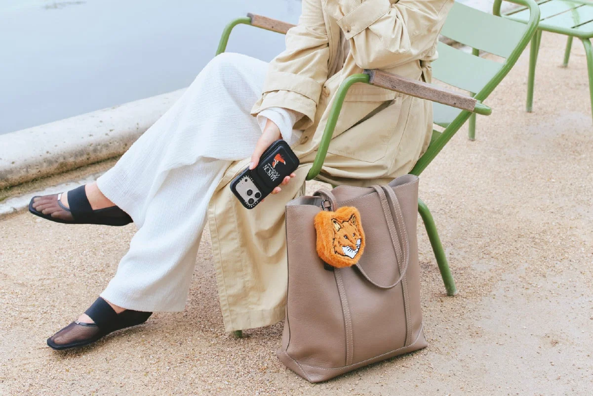 屋外の椅子に座っている女性が、ベージュのトレンチコートと白いパンツを着用し、スマートフォンとキツネのチャームが付いたトートバッグを持っている様子。リラックスした雰囲気のファッションスナップです。