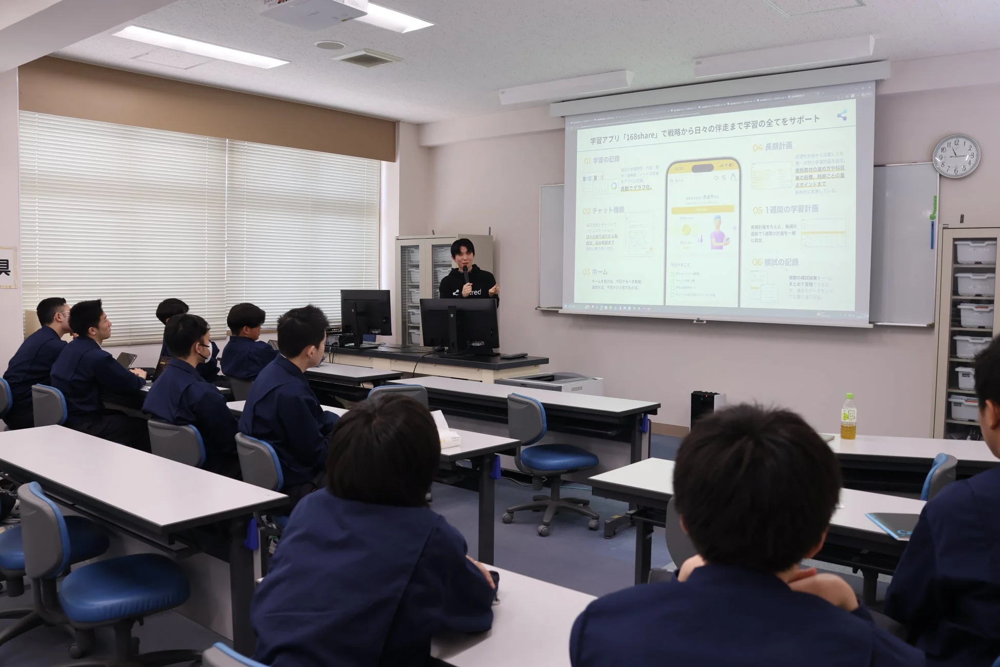 教室で講師が学習アプリ「168share」の機能（学習記録、チャット、計画、模試記録など）を学生にプレゼンしています。学生たちは熱心に画面を見つめ、デジタル学習支援ツールについて学んでいます。