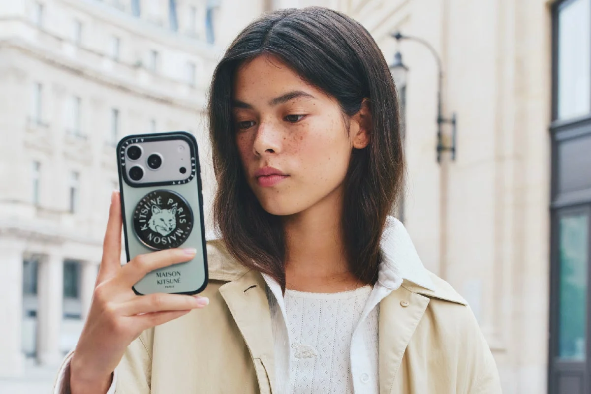 街中でスマートフォンを構える女性と、その奥に立つ男性のポートレート。女性はカメラ目線で、キツネのデザインが施されたスマホケースが印象的です。