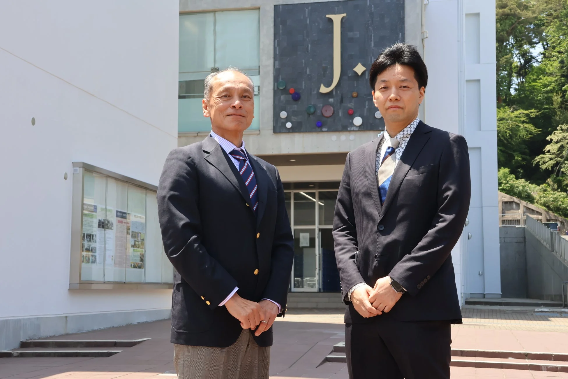 スーツ姿の男性二人が、大きな「J.」のロゴとカラフルな装飾が施された建物の前に立っています。プロフェッショナルな雰囲気で、晴れた日の屋外での一枚です。