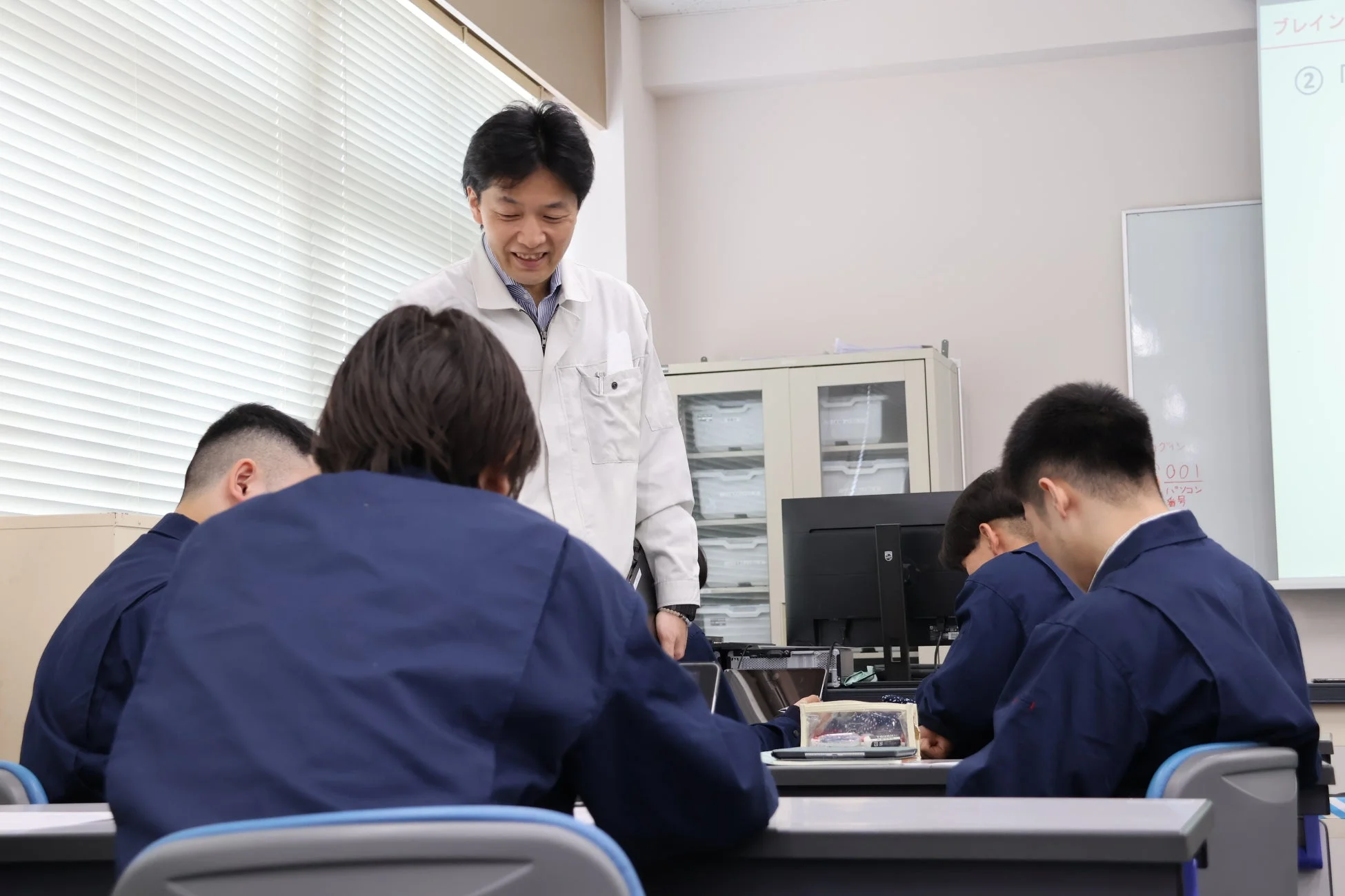 白い作業服を着た講師が、青い作業服の生徒たちが机でパソコンを使った学習や実習に取り組む様子を笑顔で見守っています。和やかな雰囲気の中で真剣に学んでいる様子が伺えます。