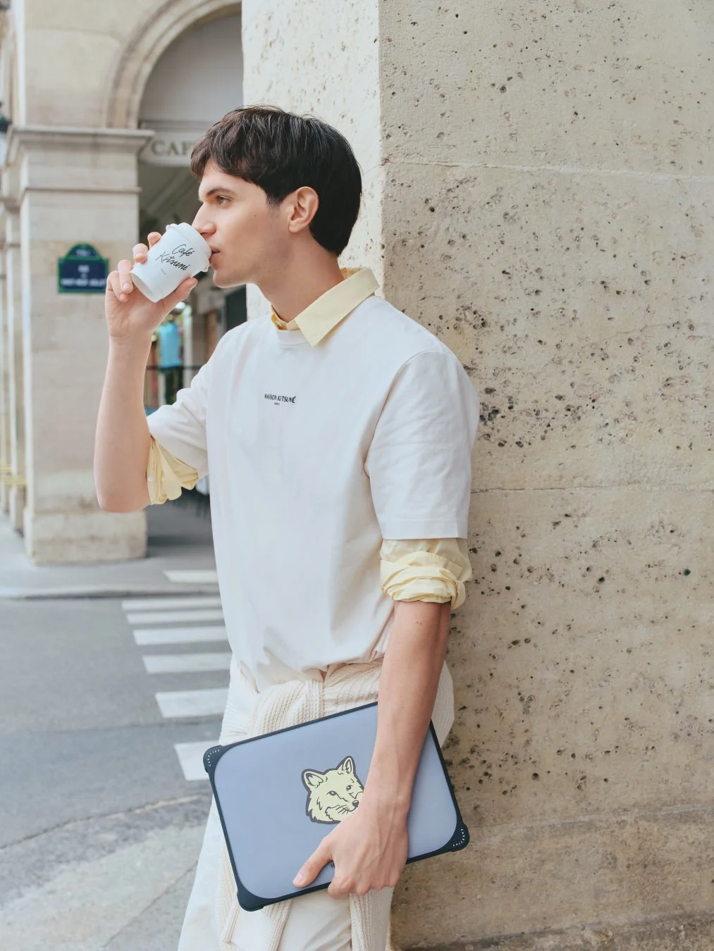 街角でコーヒーを飲む男性。メゾンキツネのシャツとキツネのロゴ入りノートパソコンケースが目を引きます。
