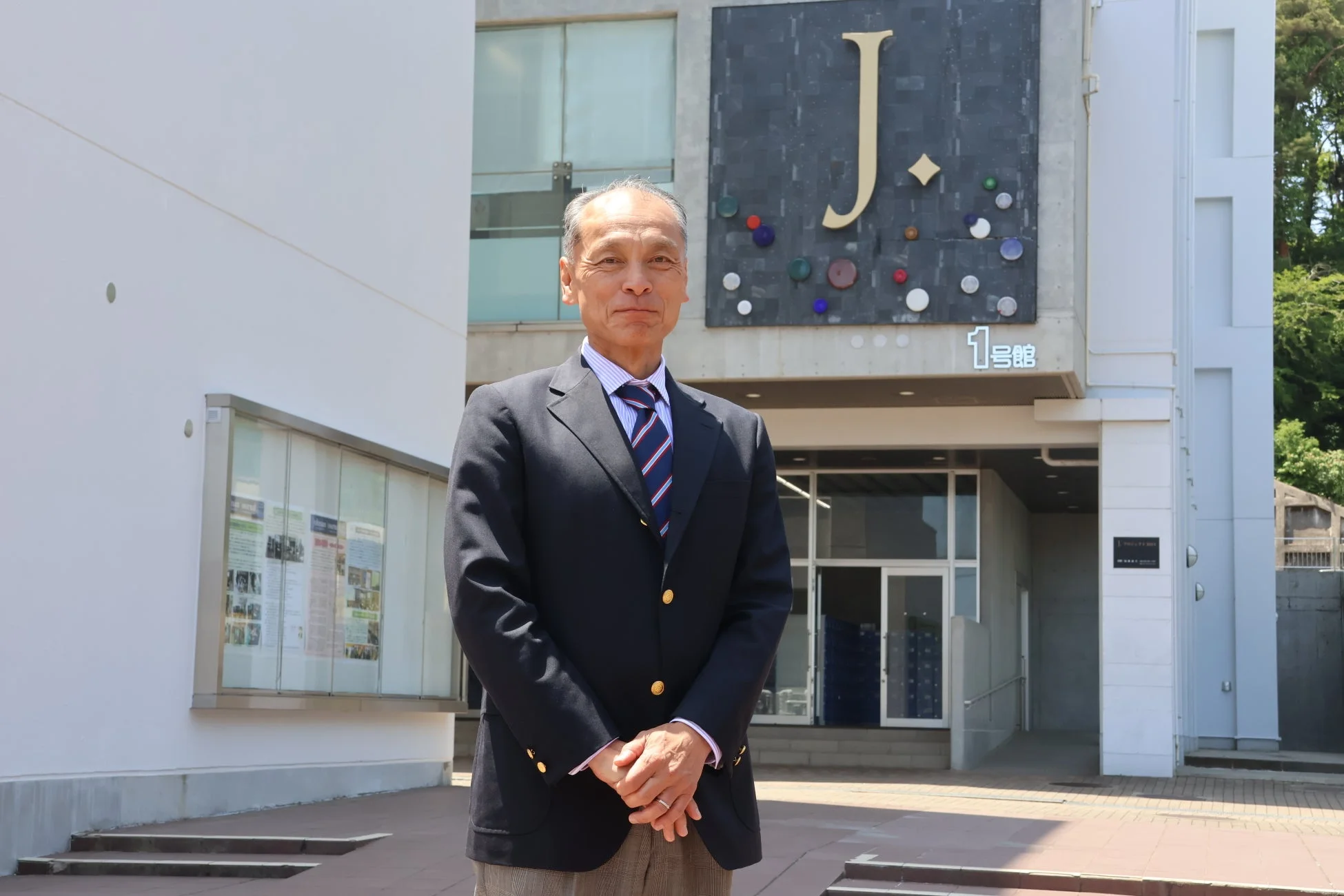 スーツを着た年配の男性が、Jのロゴと「1号館」の表示があるモダンな建物の前で立っています。日中の屋外で、大学のような教育機関のキャンパス風景と見られます。