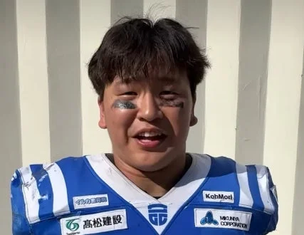 青いアメフトユニフォームを着た男性が、目の下にアイブラックを施し、屋外で笑顔を見せています。