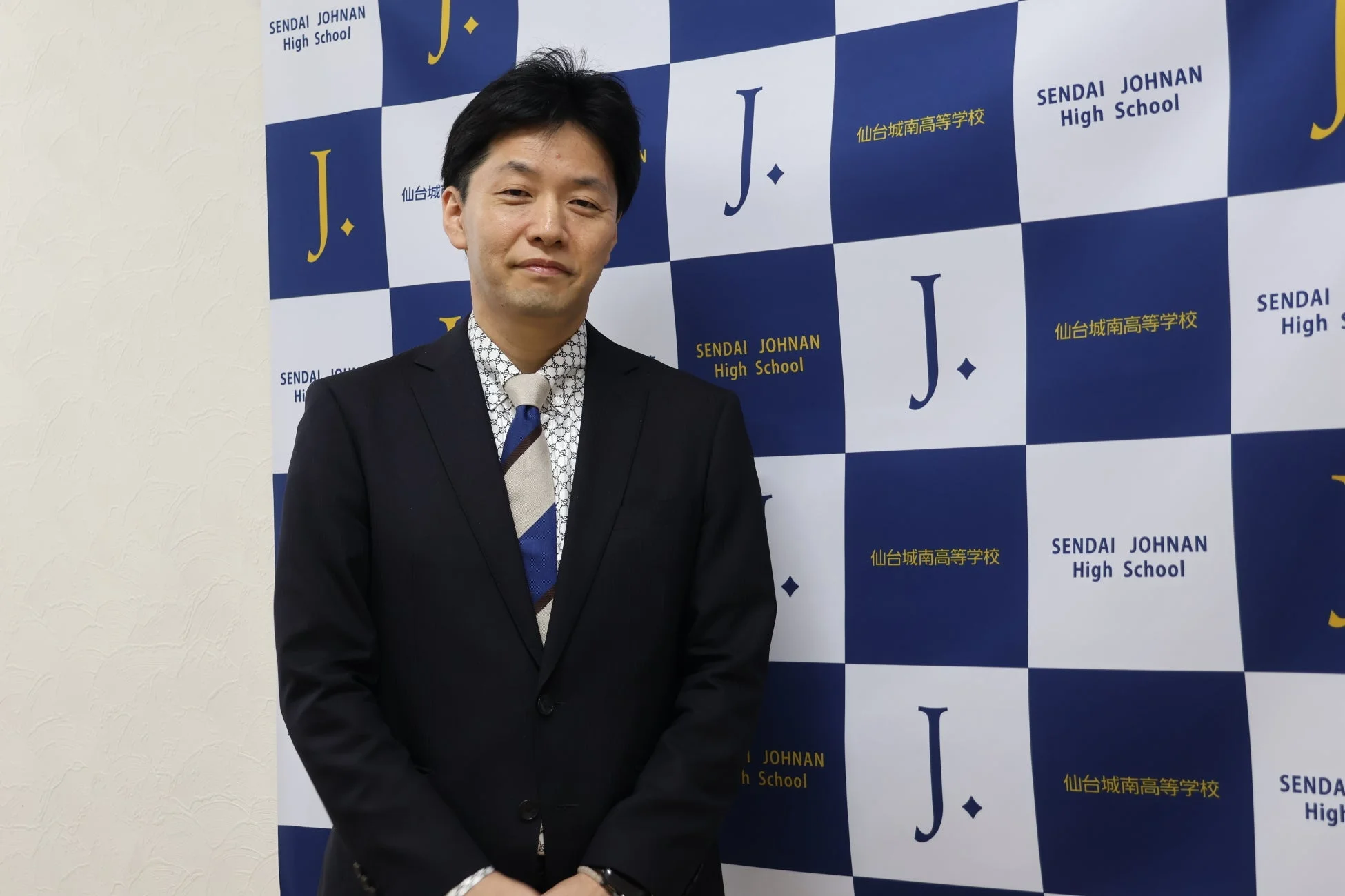 スーツを着用した男性が、仙台城南高等学校のロゴと校名がデザインされた背景の前で立っている。真面目な表情でカメラを見つめており、公式なイベントや発表の場を思わせる。